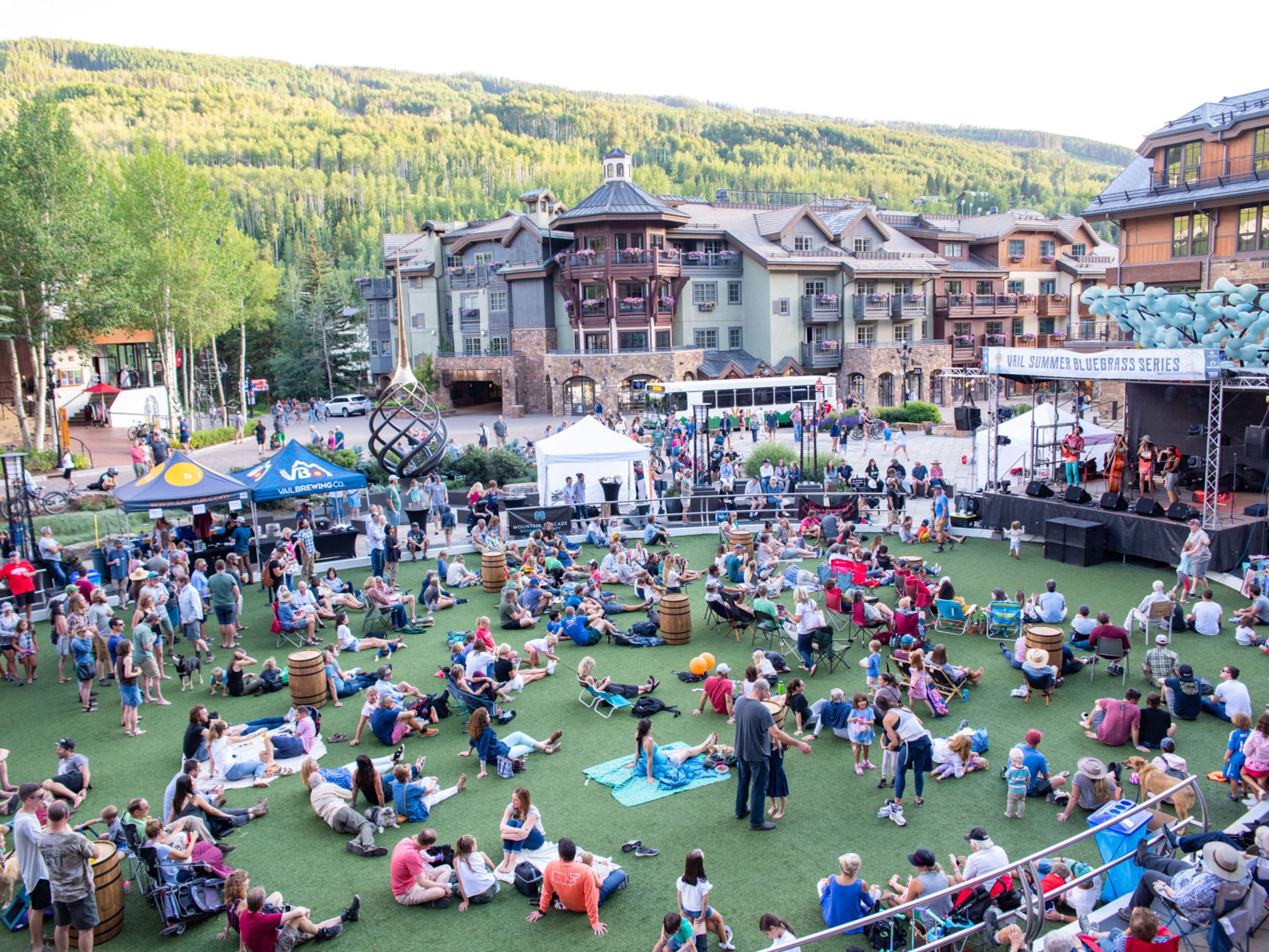 A large outdoor event with people sitting on grass, a stage with performers, and a backdrop of buildings and mountains.