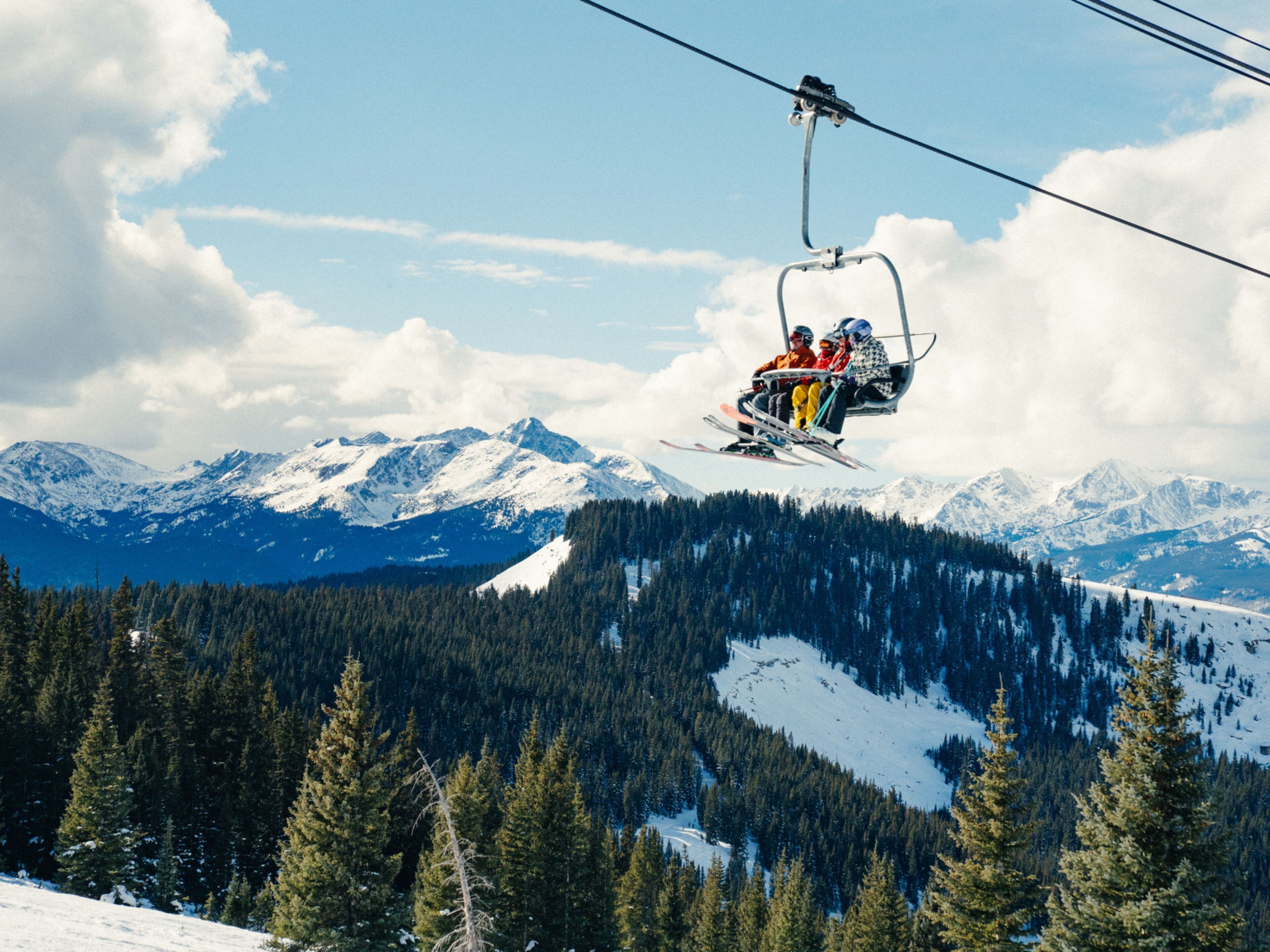 A ski lift with passengers, snow-covered mountains, and a forested valley.