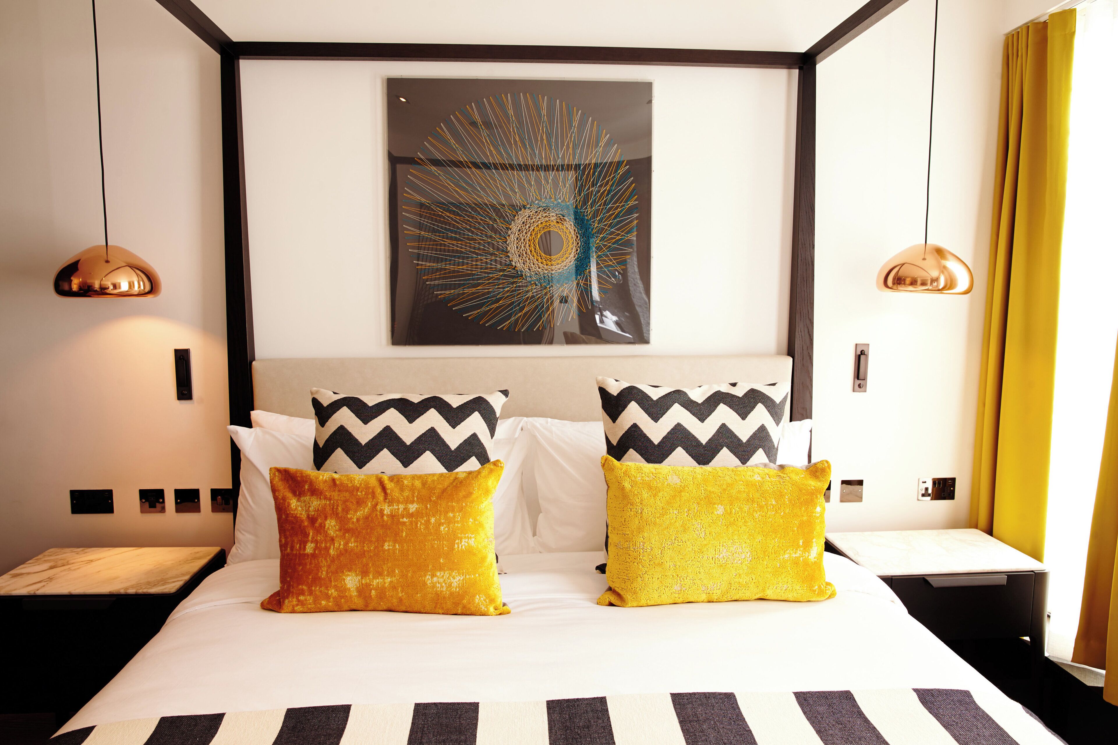 Habitación de hotel con cama, dos almohadas, una obra de arte en la pared y dos lámparas colgantes.