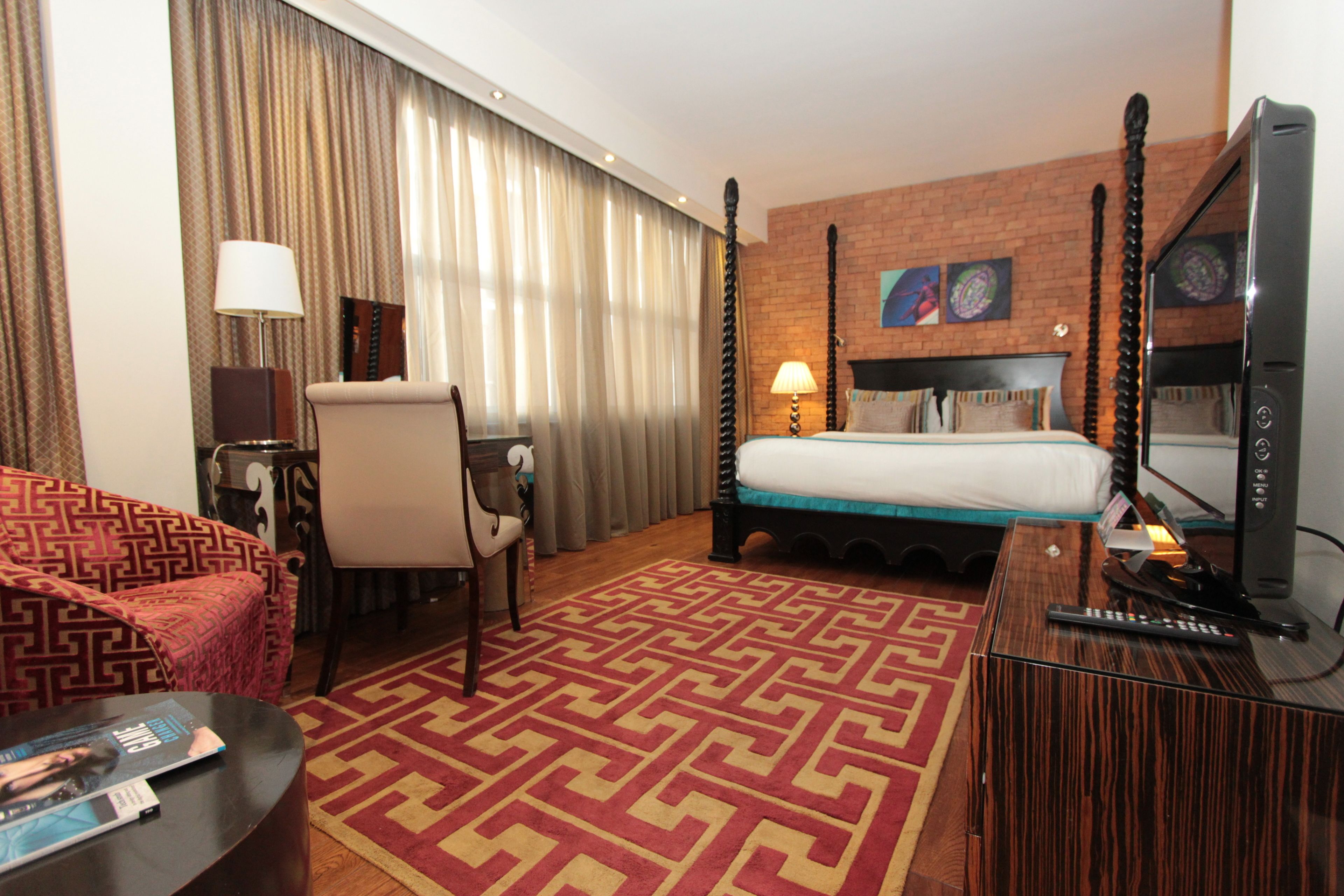 A hotel room with a four-poster bed, a TV, a desk with a lamp, a patterned sofa, and a red and beige rug.