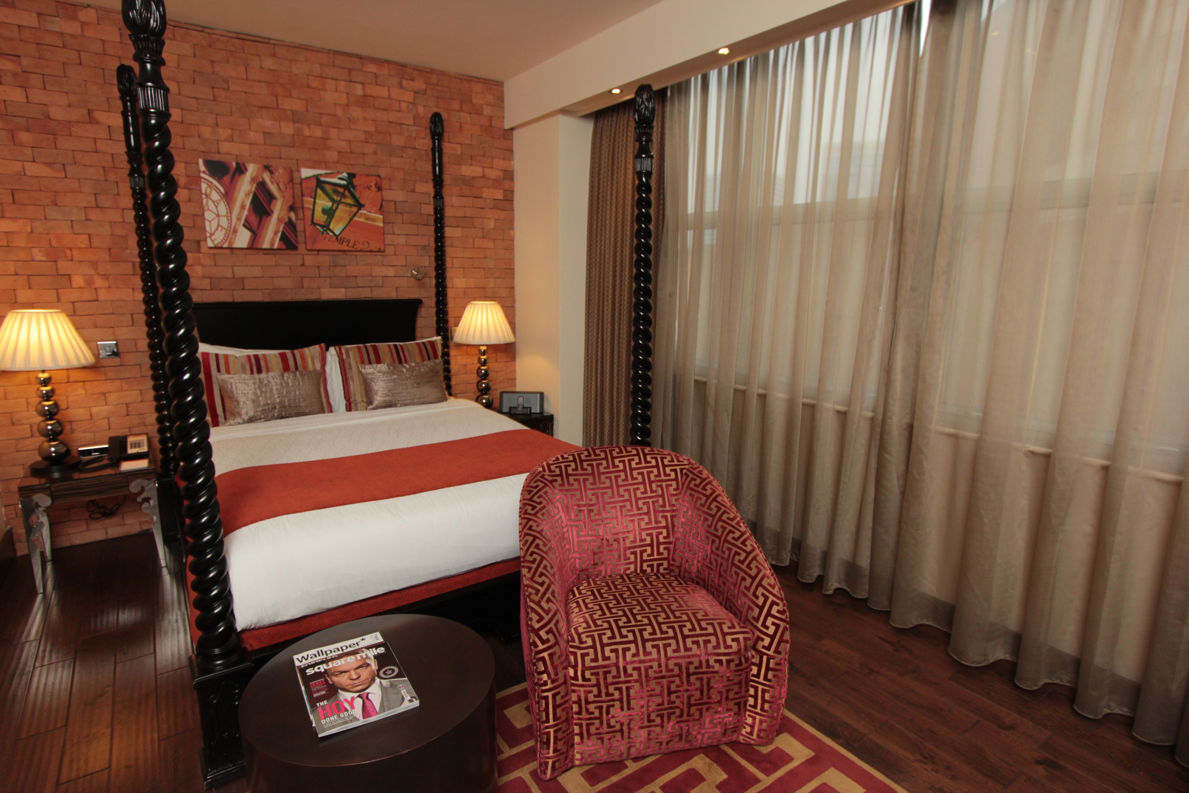 A hotel room with a four-poster bed, a patterned armchair, a round table with a magazine, and a brick wall with artwork.