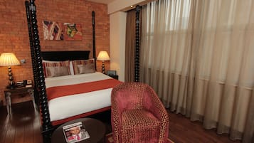 A hotel room with a four-poster bed, a patterned armchair, a round table with a magazine, and a brick wall with artwork.
