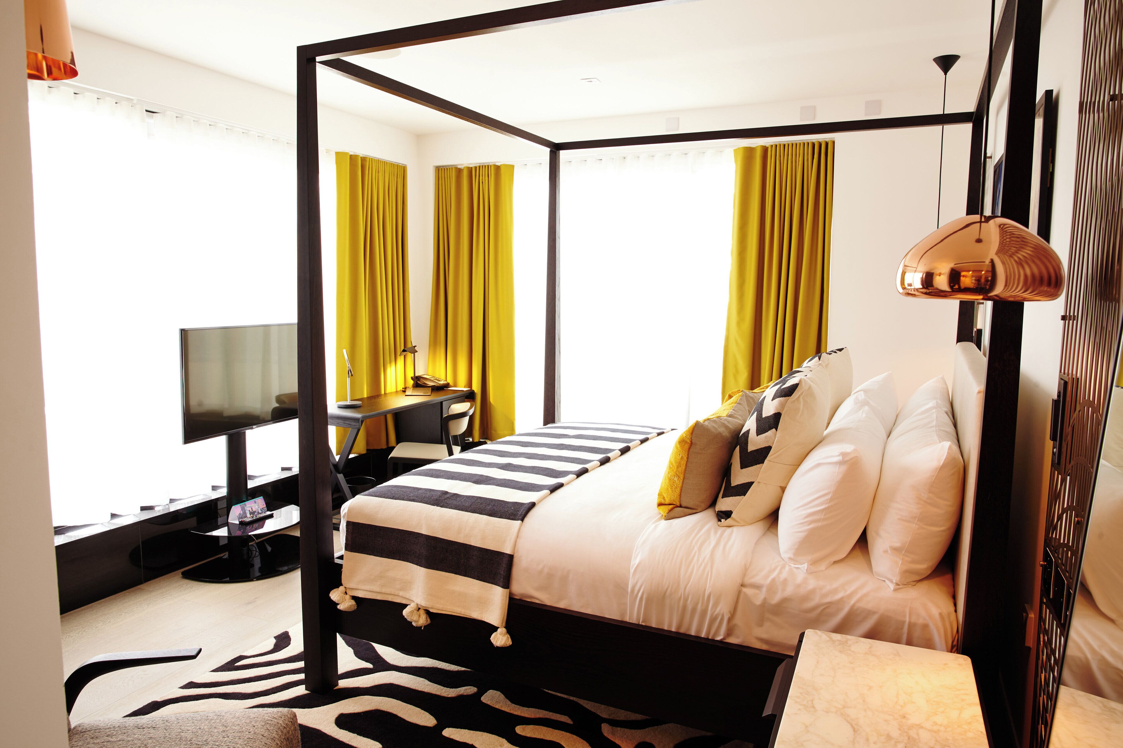 Una cama con dosel, un tapete con estampado de cebra, un escritorio con una silla, un televisor y cortinas amarillas.