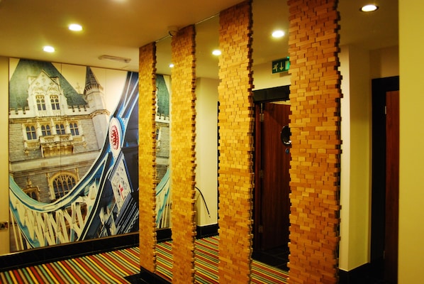 Un couloir orné d’une fresque représentant un bâtiment, avec un escalier mécanique, des colonnes en briques et un tapis à rayures.