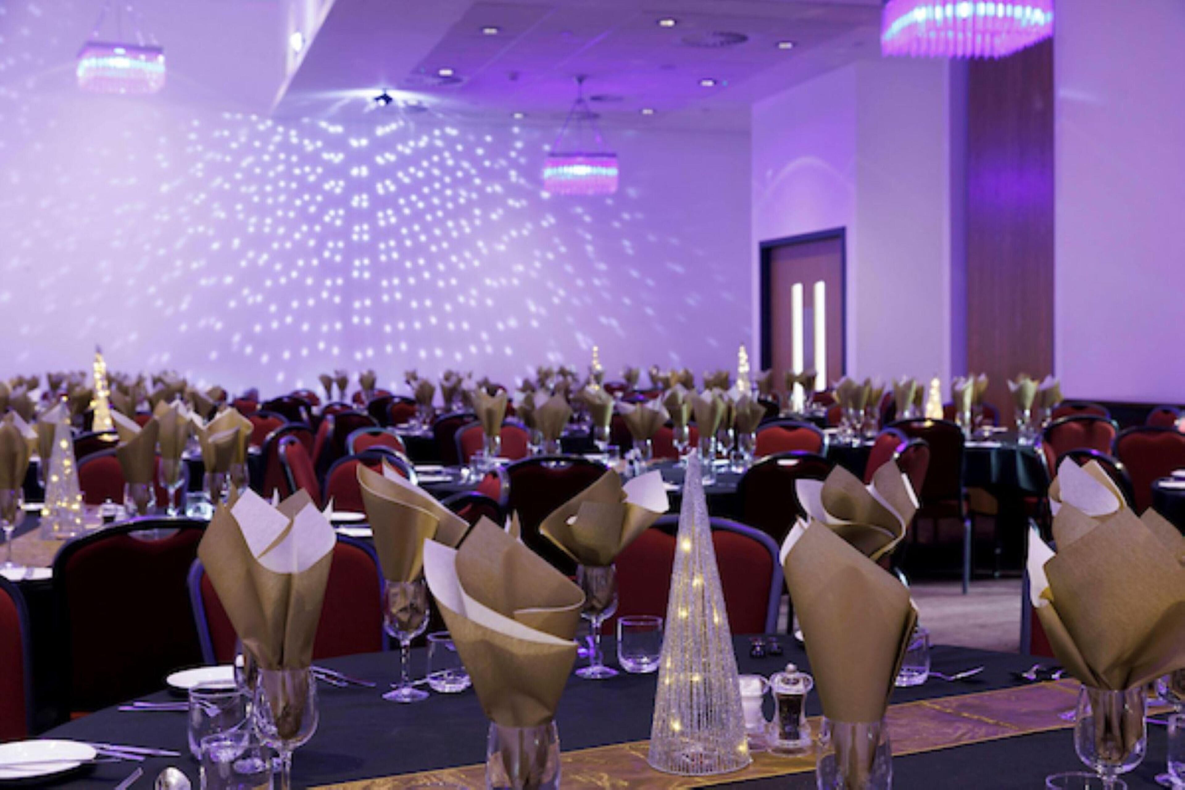 A banquet hall with tables set for a formal event, featuring gold napkin holders and clear glassware.
