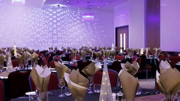A banquet hall with tables set for a formal event, featuring gold napkin holders and clear glassware.