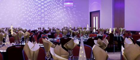 A banquet hall with tables set for a formal event, featuring gold napkin holders and clear glassware.