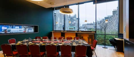 A dining area with a large table set for a meal, red chairs, and a view of buildings through large windows.