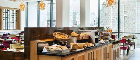 Un espace buffet pour le petit-déjeuner proposant une variété de pains et de viennoiseries, une machine à café et des tables où s’asseoir.