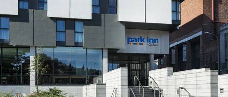 Un edificio moderno con el letrero 'Park Inn by Radisson', que presenta una escalera que lleva a la entrada, ventanas amplias y una sección de ladrillo a la derecha.