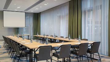 Un salón de conferencias con una mesa larga, sillas, una pantalla de proyector y un amplio ventanal con cortinas.