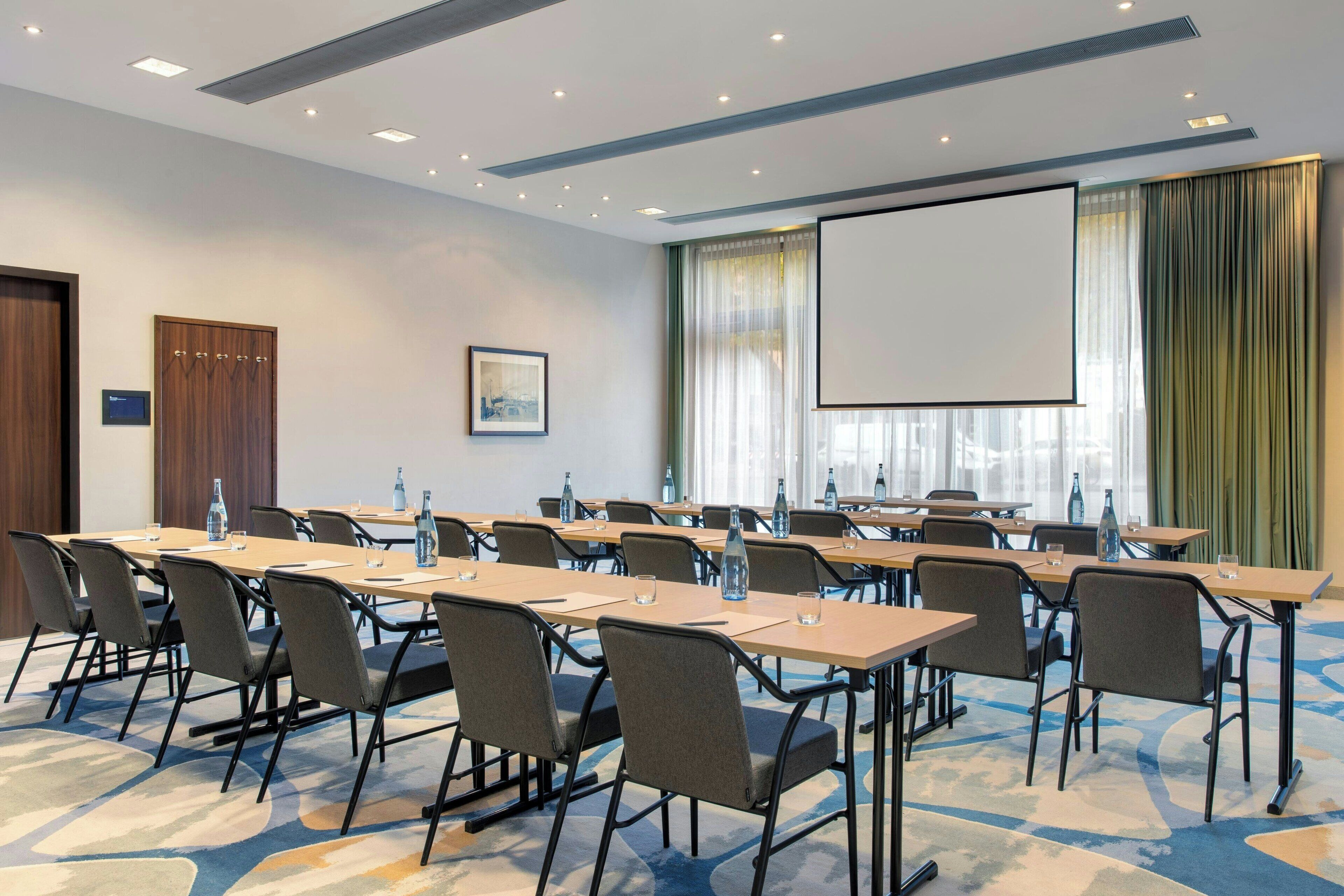 Un salón de conferencias con varias mesas, sillas, una pantalla de proyector y un ventanal con cortinas.