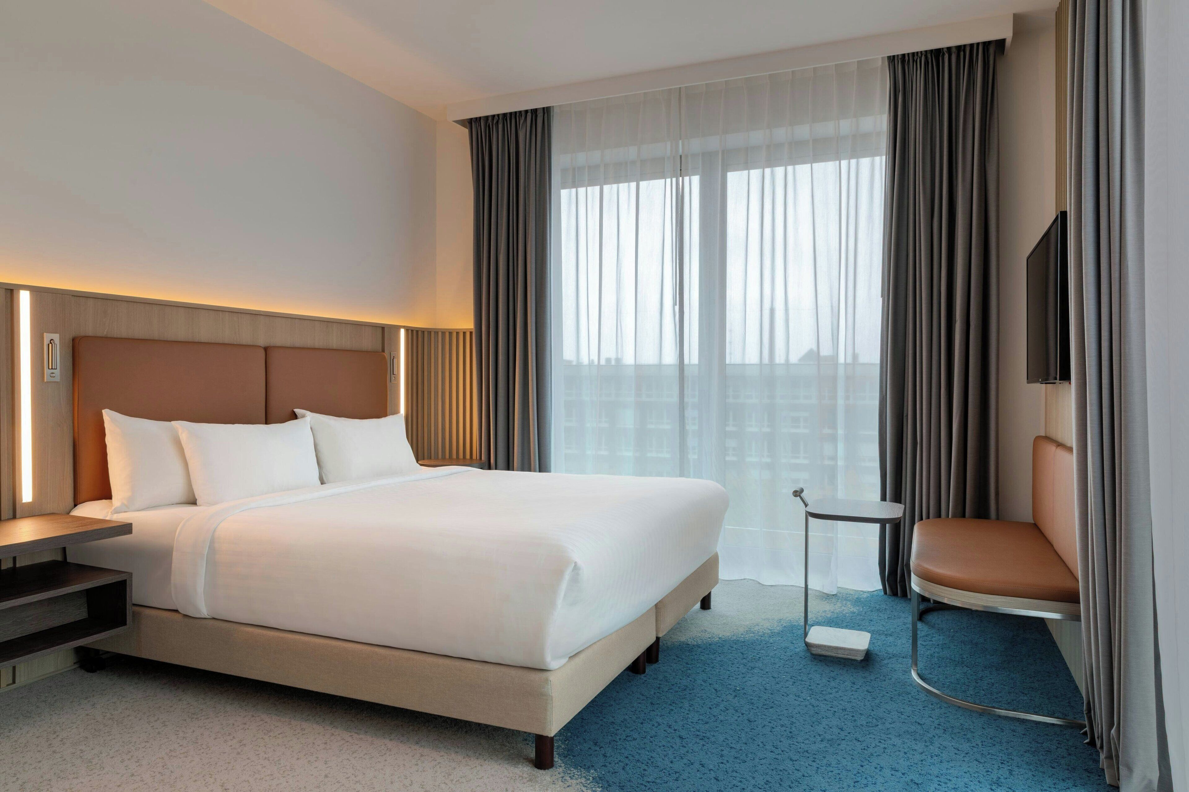 A hotel room with a large bed, a bedside table, a chair, a TV, and a window with curtains.