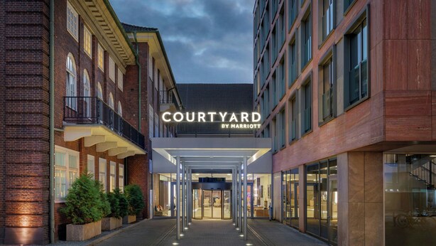 Entrada de un hotel con un letrero que dice "COURTYARD BY MARRIOTT", con un diseño moderno, puertas de vidrio y fachada de ladrillo.