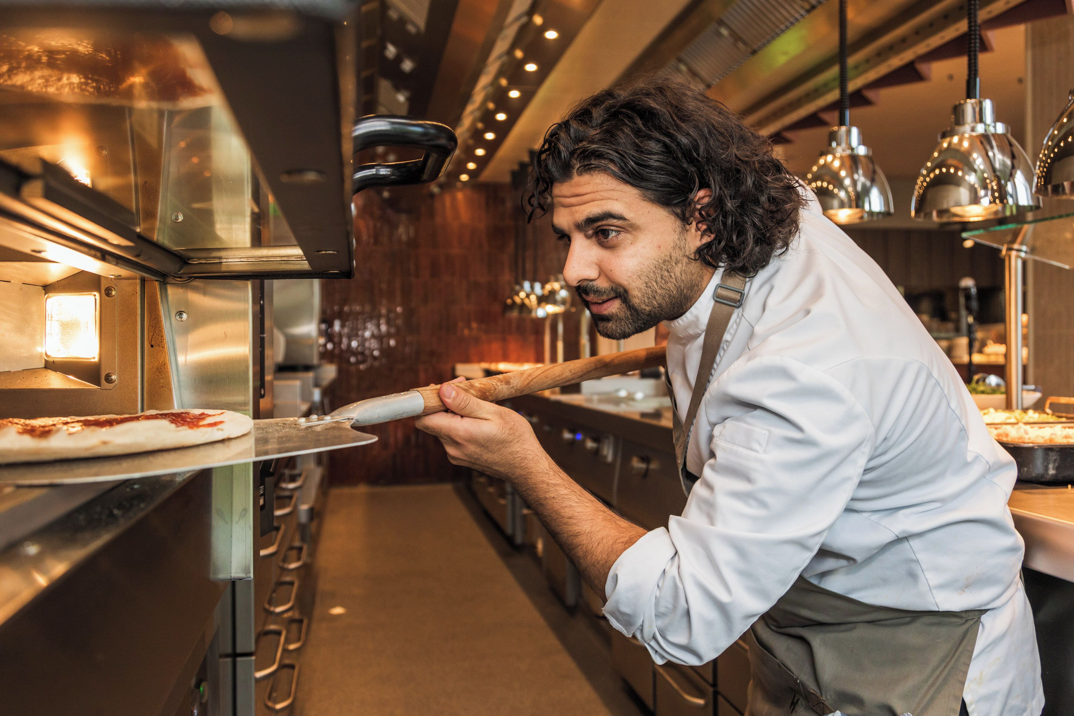 Une cuisine équipée d’appareils électroménagers en acier inoxydable, une personne utilisant une pelle à pizza et un éclairage au plafond.