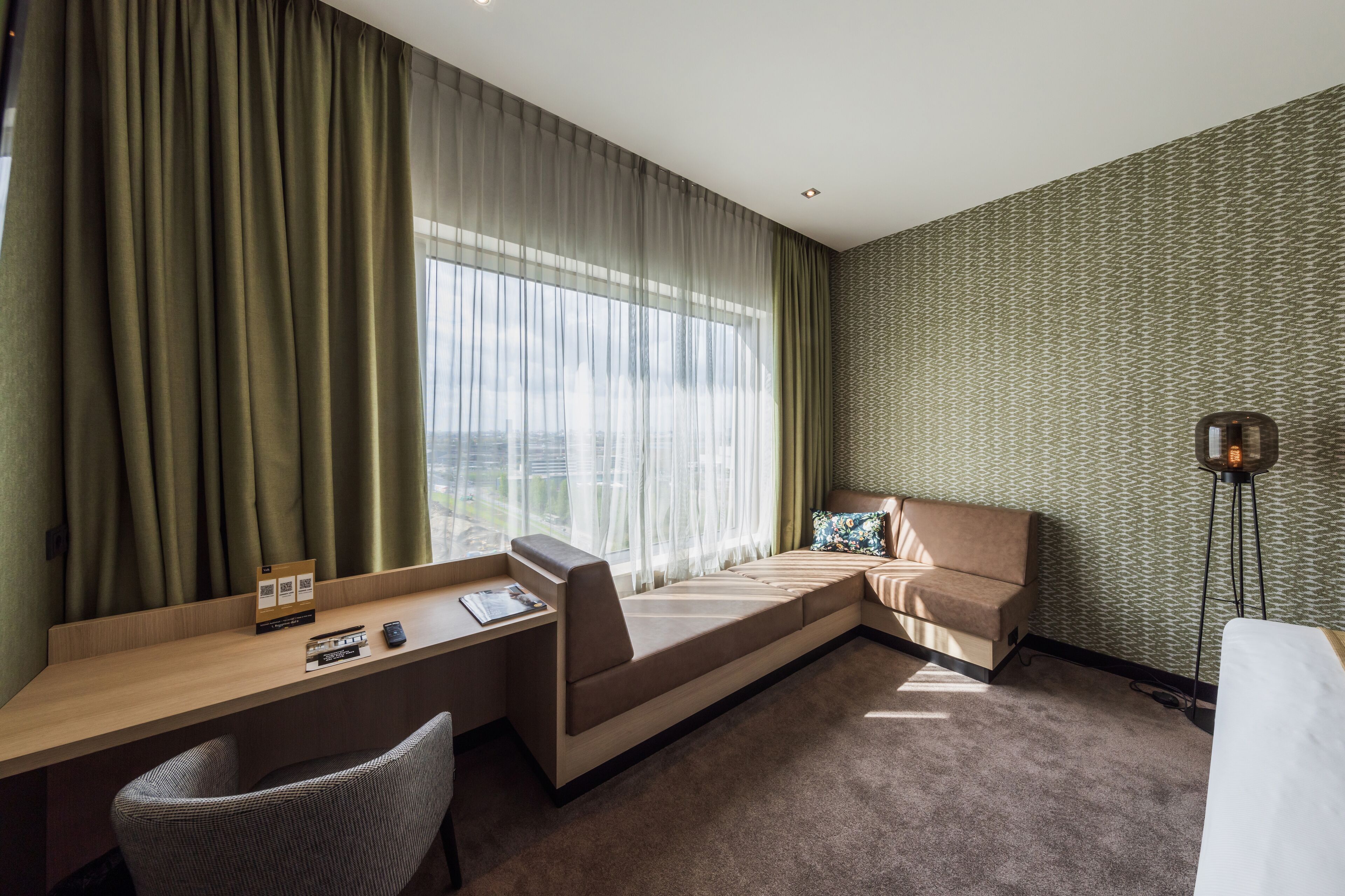 Habitación de hotel con escritorio, sofá, sillón y un ventanal con cortinas translúcidas.