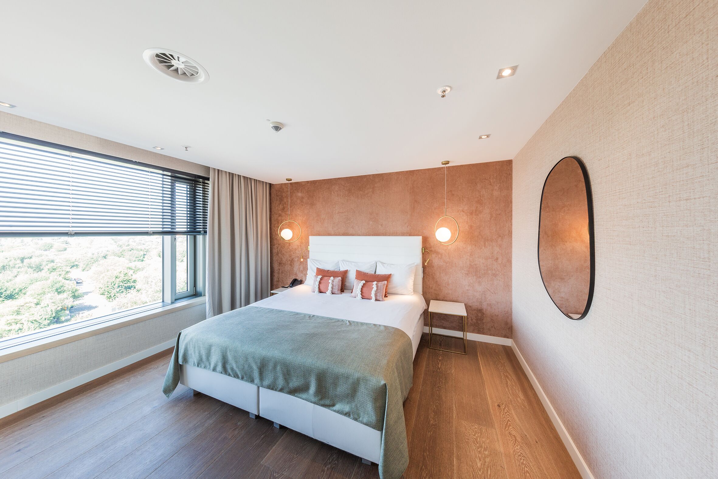 A modern bedroom with a large bed, a bedside table, a mirror, and a window with blinds.