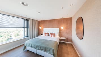 A modern bedroom with a large bed, a bedside table, a mirror, and a window with blinds.