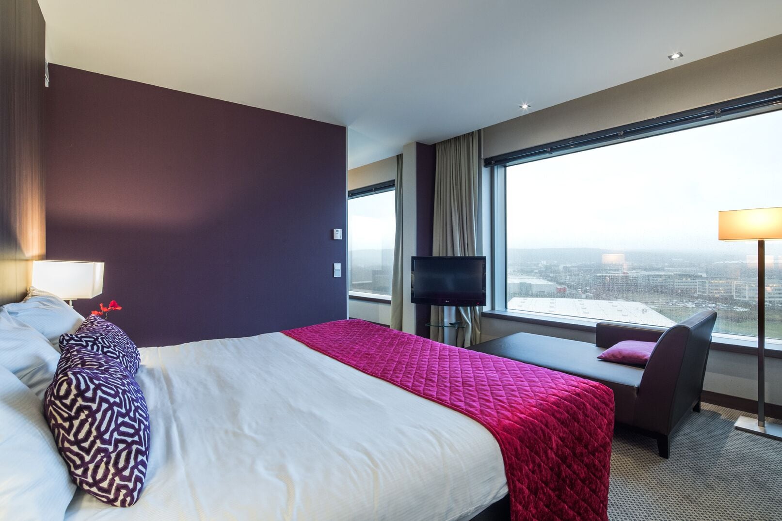Een hotelkamer met een groot bed, een bureau, een stoel, een televisie en uitzicht op de stad.
