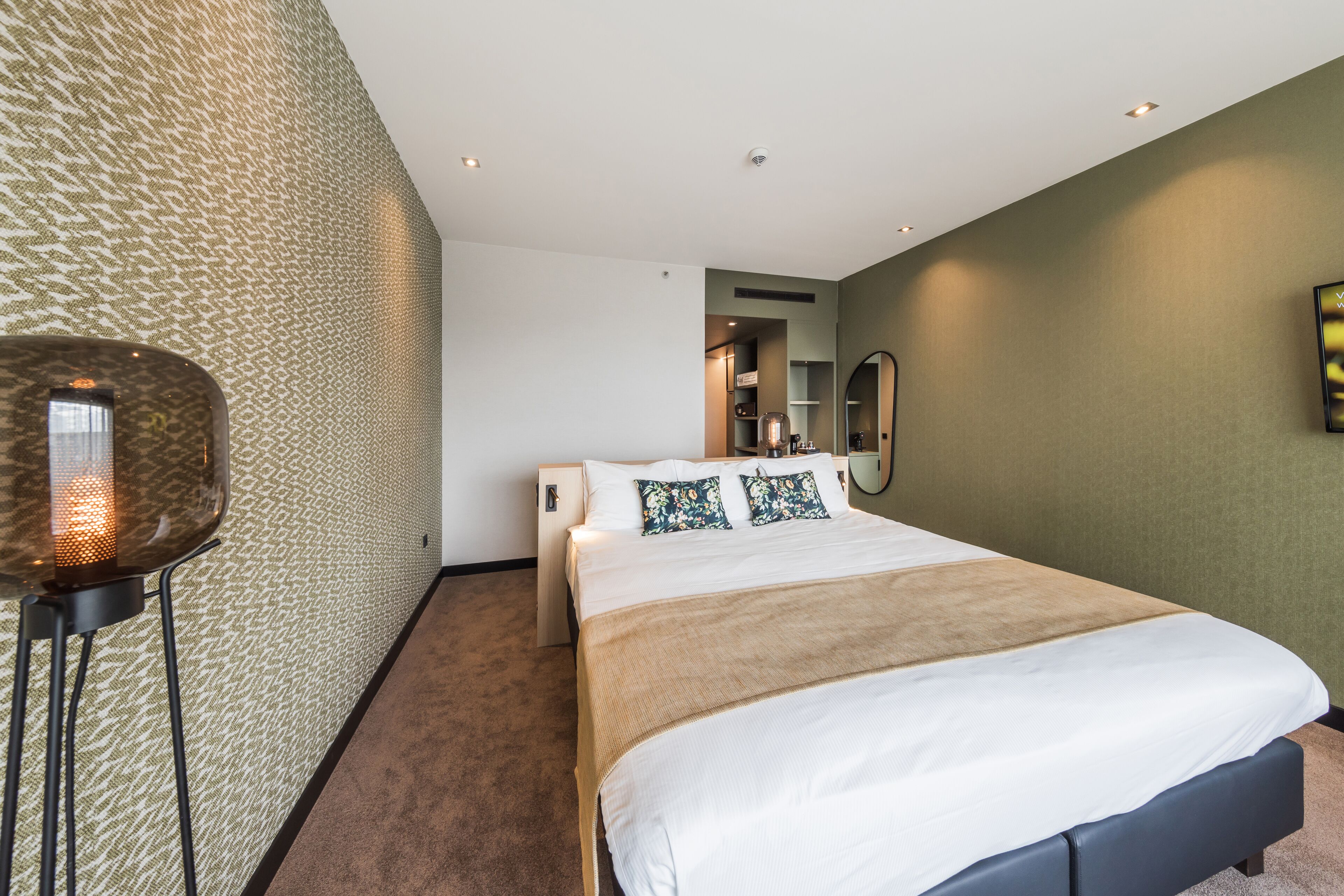 A modern hotel room with a large bed, a standing lamp, and a mirror on the wall.