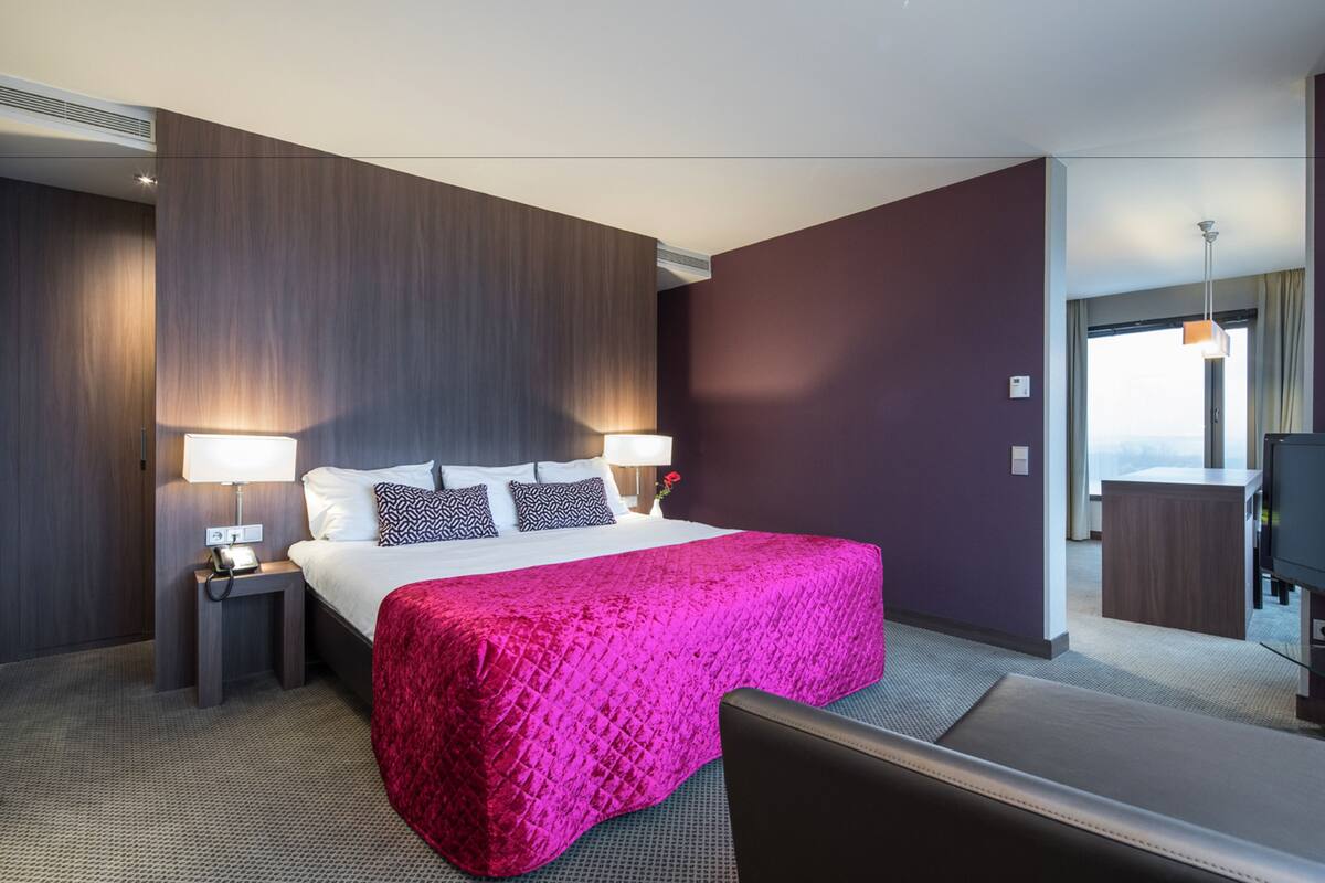 A hotel room with a large bed, two bedside tables with lamps, a flat-screen TV, and a view of the city.