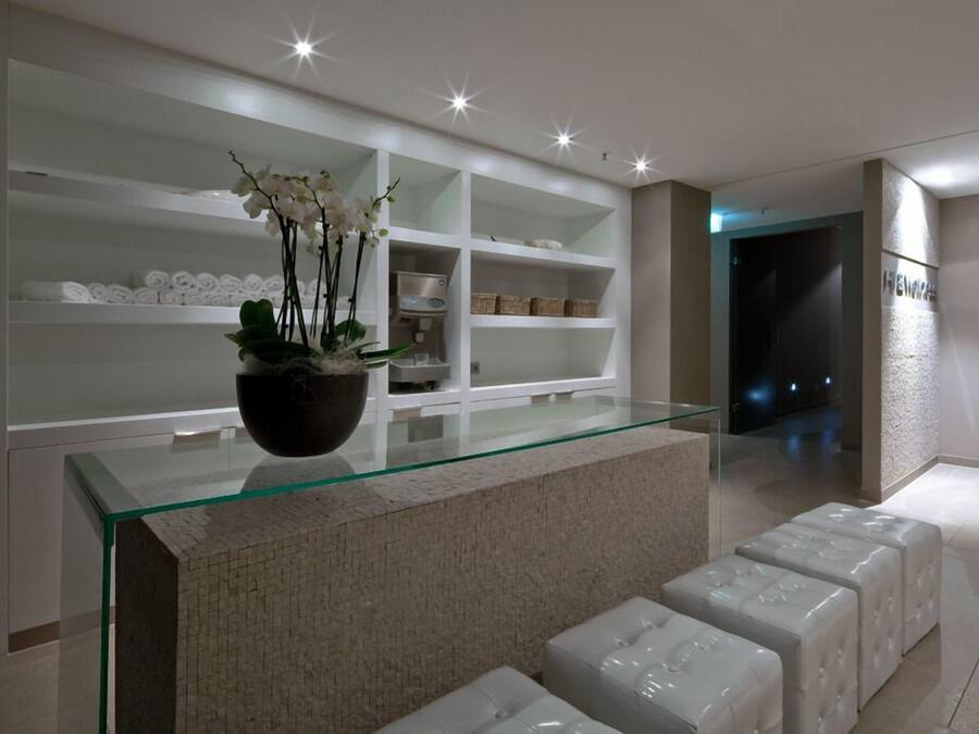 A modern hotel lobby with a glass reception desk, white shelves with towels, and a potted plant.