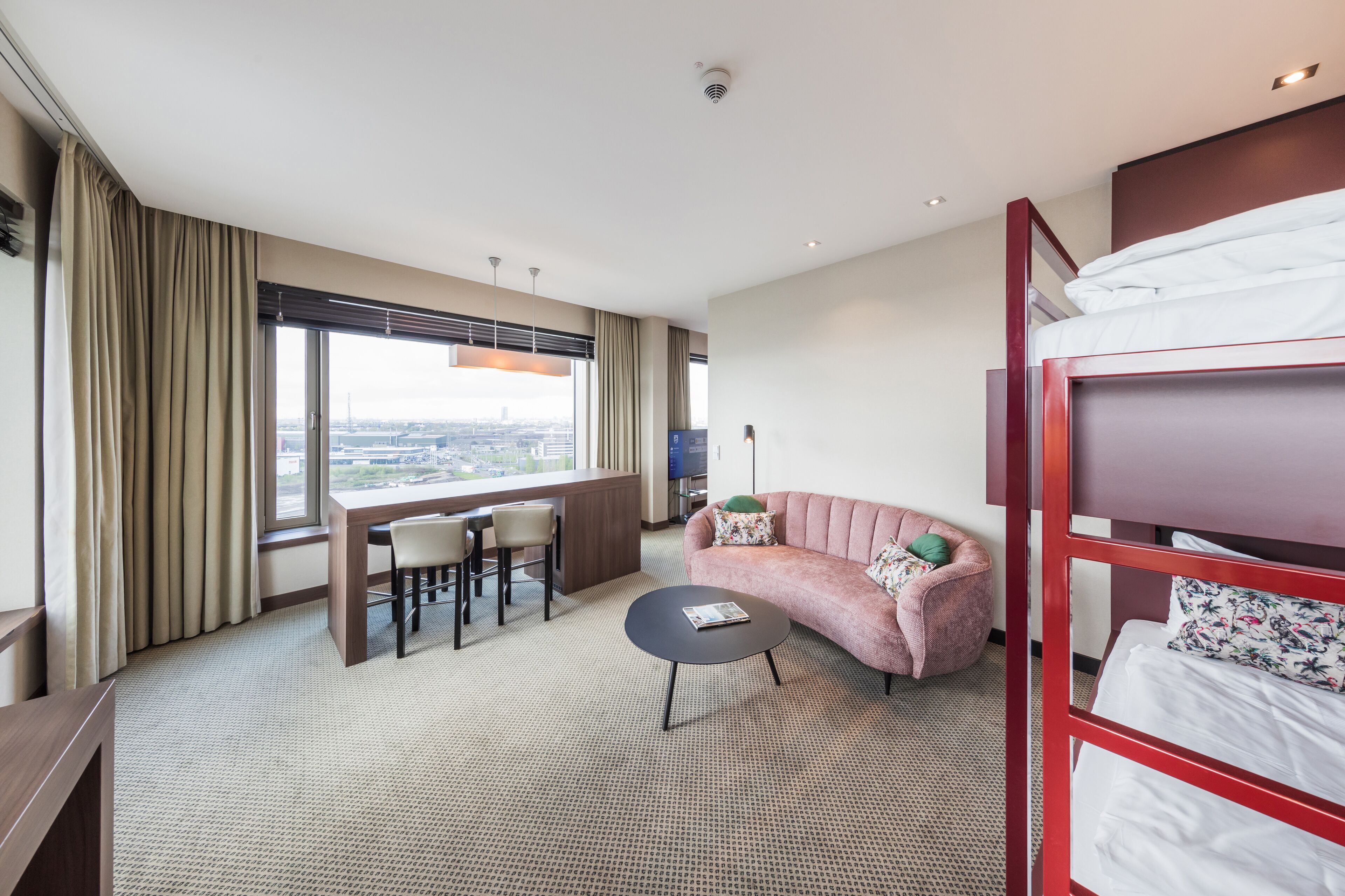 Ein Hotelzimmer mit Etagenbett, einer Couch, einem kleinen runden Tisch, einem Schreibtisch mit Stühlen und einem großen Fenster mit Blick auf die Stadt.