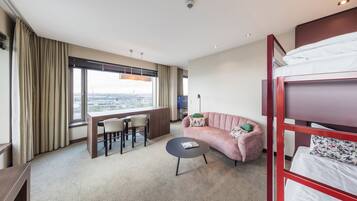 Ein Hotelzimmer mit Etagenbett, einer Couch, einem kleinen runden Tisch, einem Schreibtisch mit Stühlen und einem großen Fenster mit Blick auf die Stadt.
