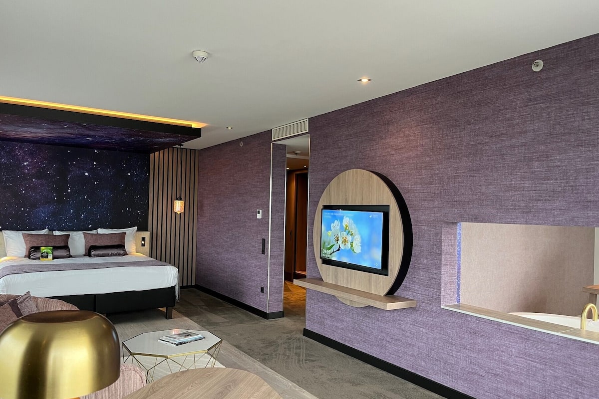 A modern hotel room with a bed, a TV mounted on the wall, and a night sky mural on the ceiling.