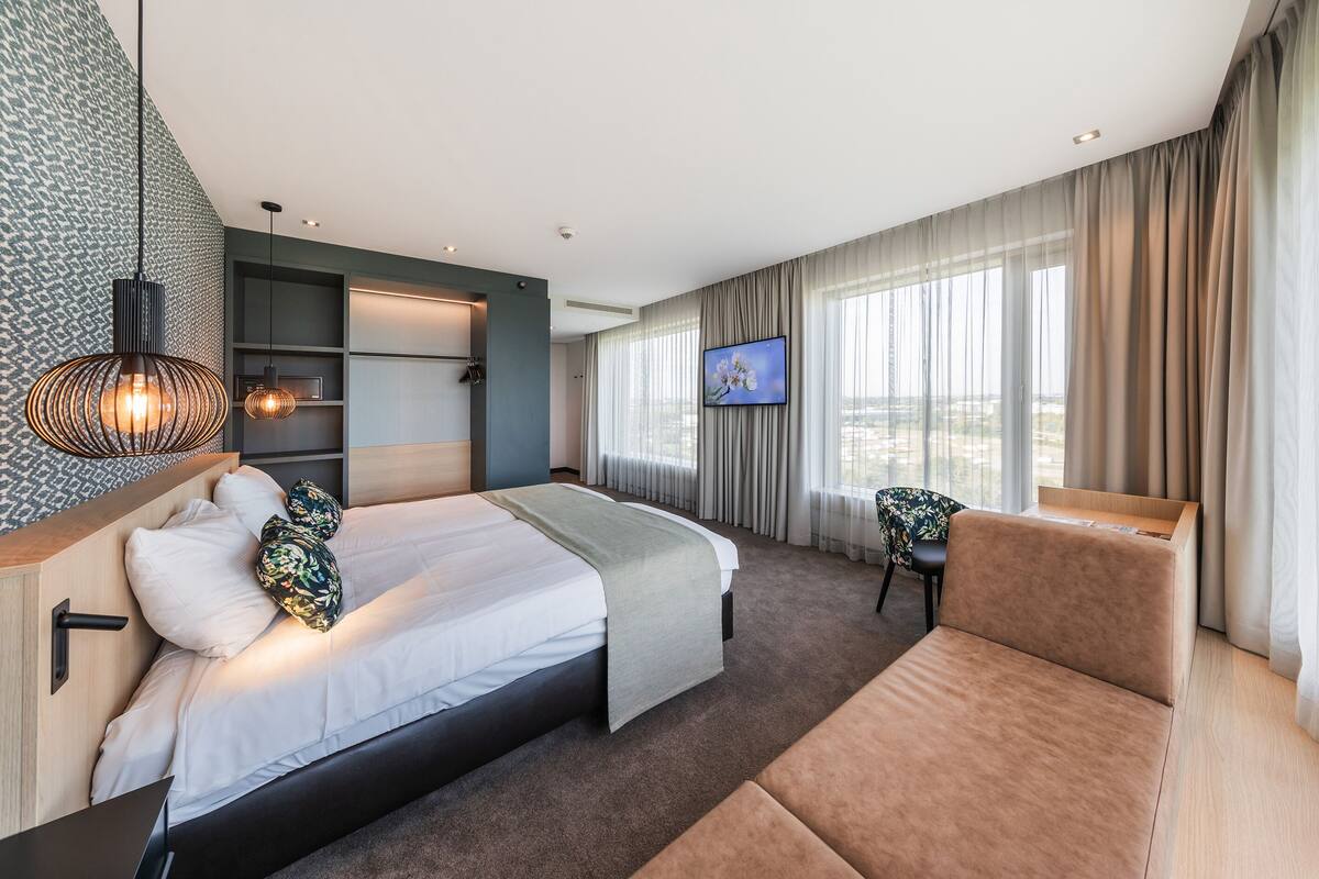 A modern hotel room with a bed, a sofa, a TV, and a city view.