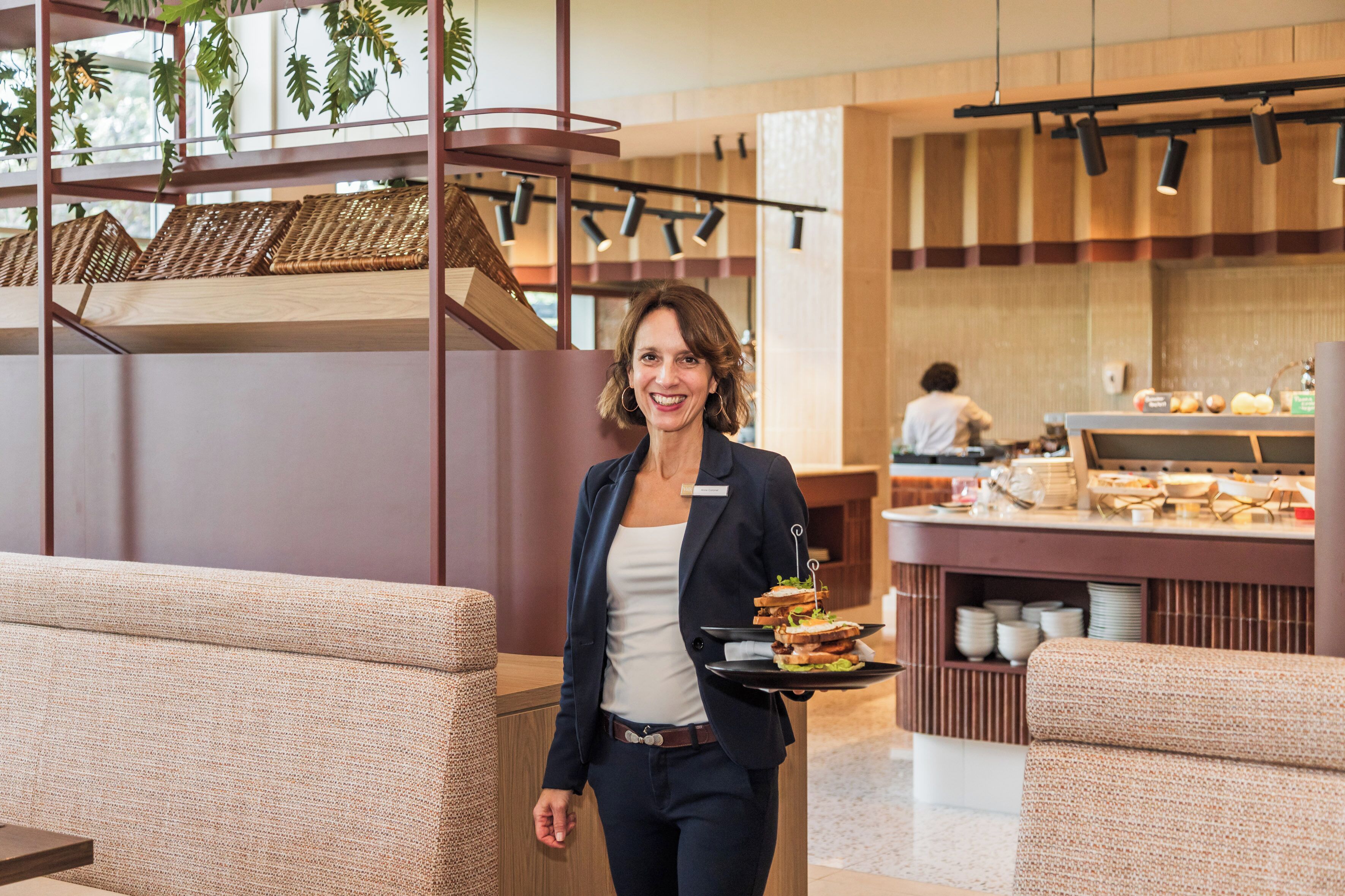 Een hotellobby met zithoeken, een buffet en een persoon die een schaal met belegde broodjes draagt.