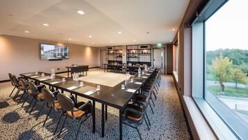 A conference room with a long table, chairs, a TV screen, and a view of a landscaped area through large windows.