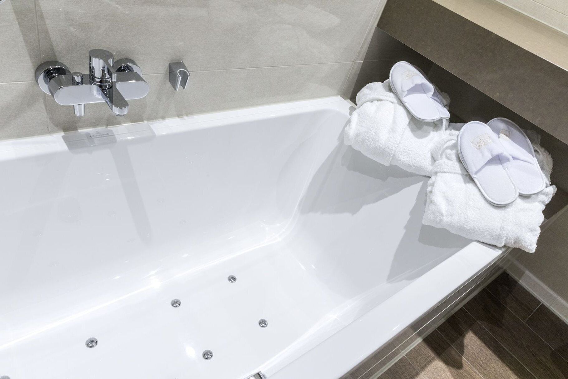 Un baño moderno con una bañera blanca, accesorios de cromo y un perchero con toallas y pantuflas blancas.