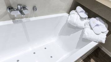 Un baño moderno con una bañera blanca, accesorios de cromo y un perchero con toallas y pantuflas blancas.