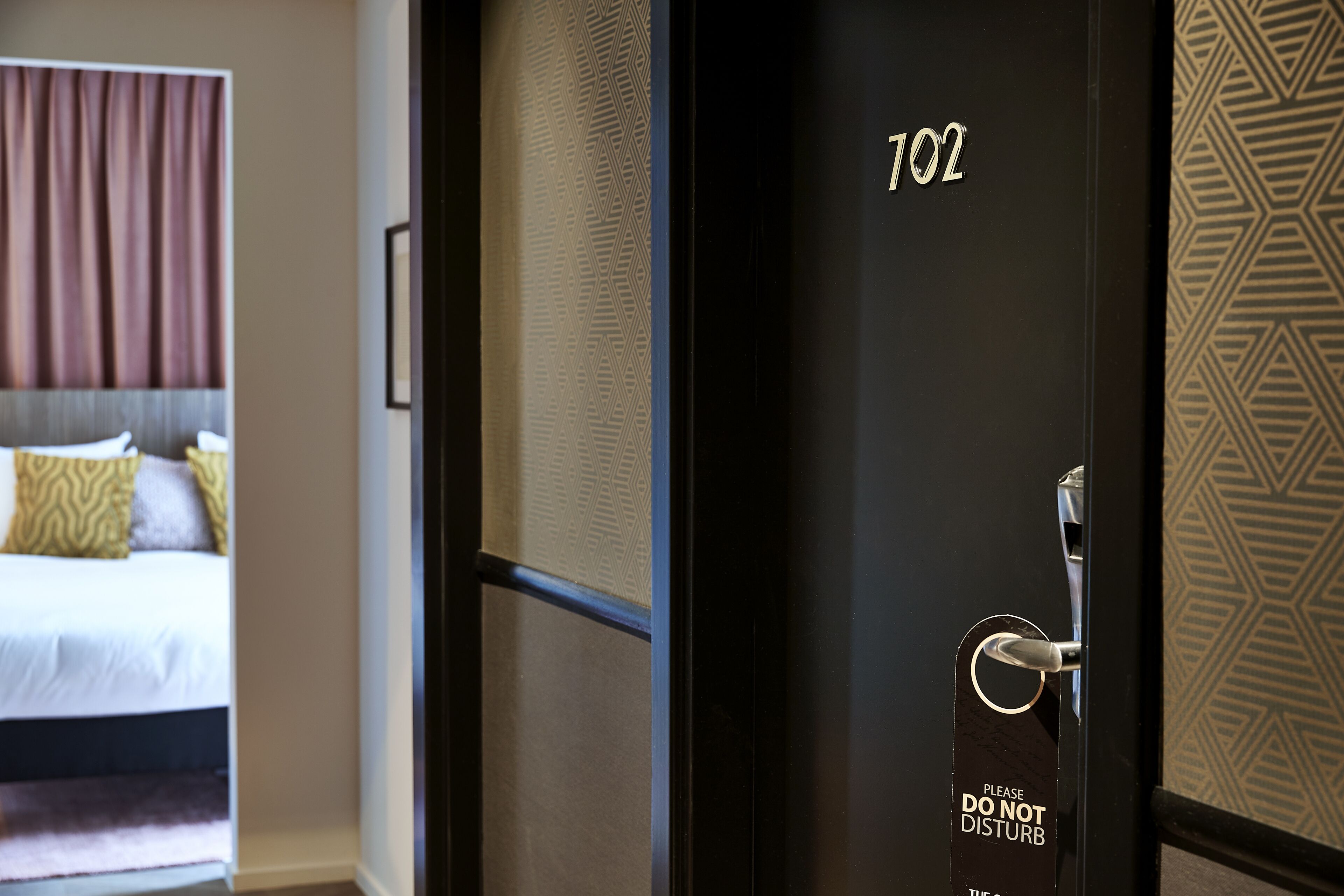 Habitación de hotel con una cama, una puerta y un cartel de 'NO MOLESTAR', además del número de habitación '102'.