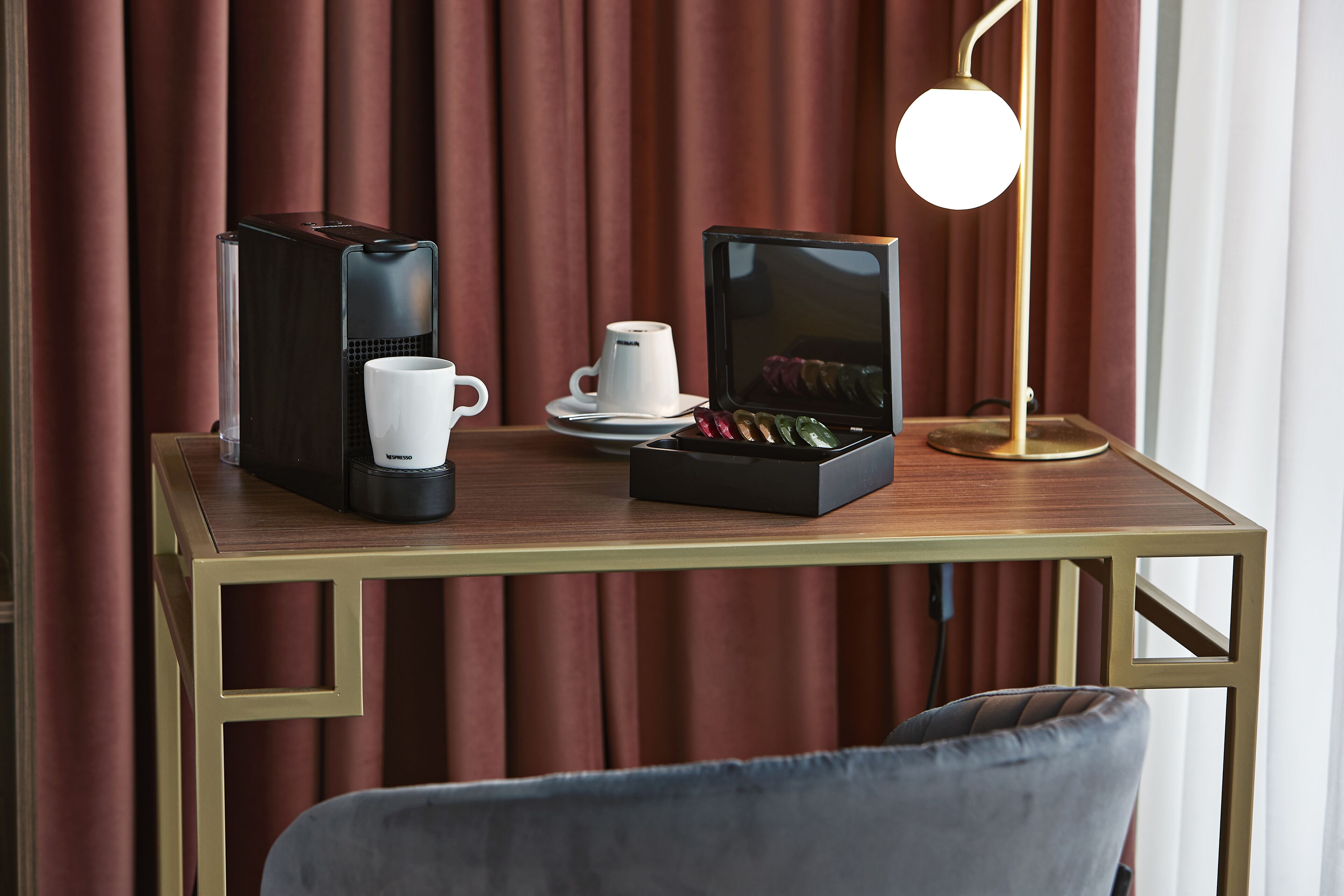 A table with a coffee maker, a cup, a box of chocolates, and a lamp.
