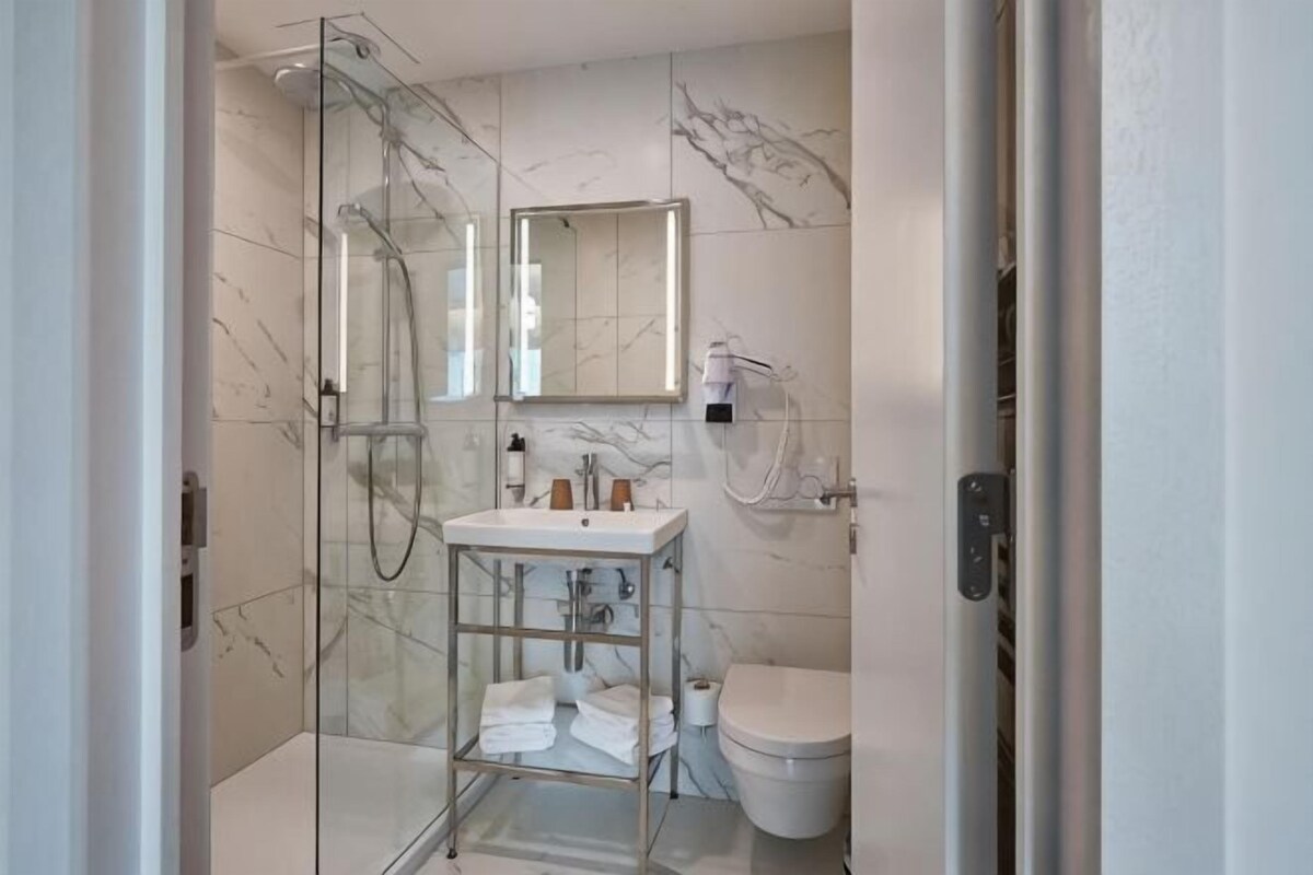 Un baño con cabina de ducha de vidrio, un lavabo blanco, un inodoro y un revestimiento de mármol en la pared.