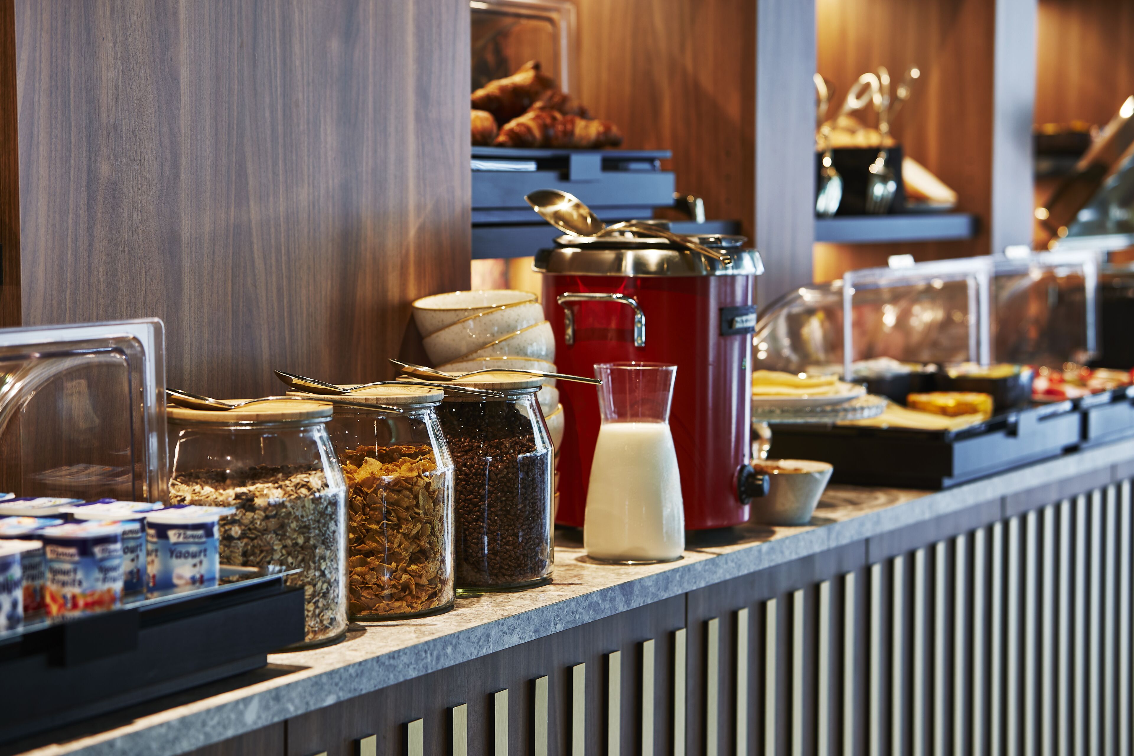 A breakfast bar with various food items and containers, including jars of granola and yogurt, a milk dispenser, and a red coffee maker.