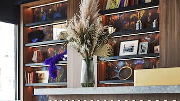 Un área de recepción con un mostrador de madera, dos taburetes rojos, un jarrón con flores secas y una estantería con libros y objetos decorativos.