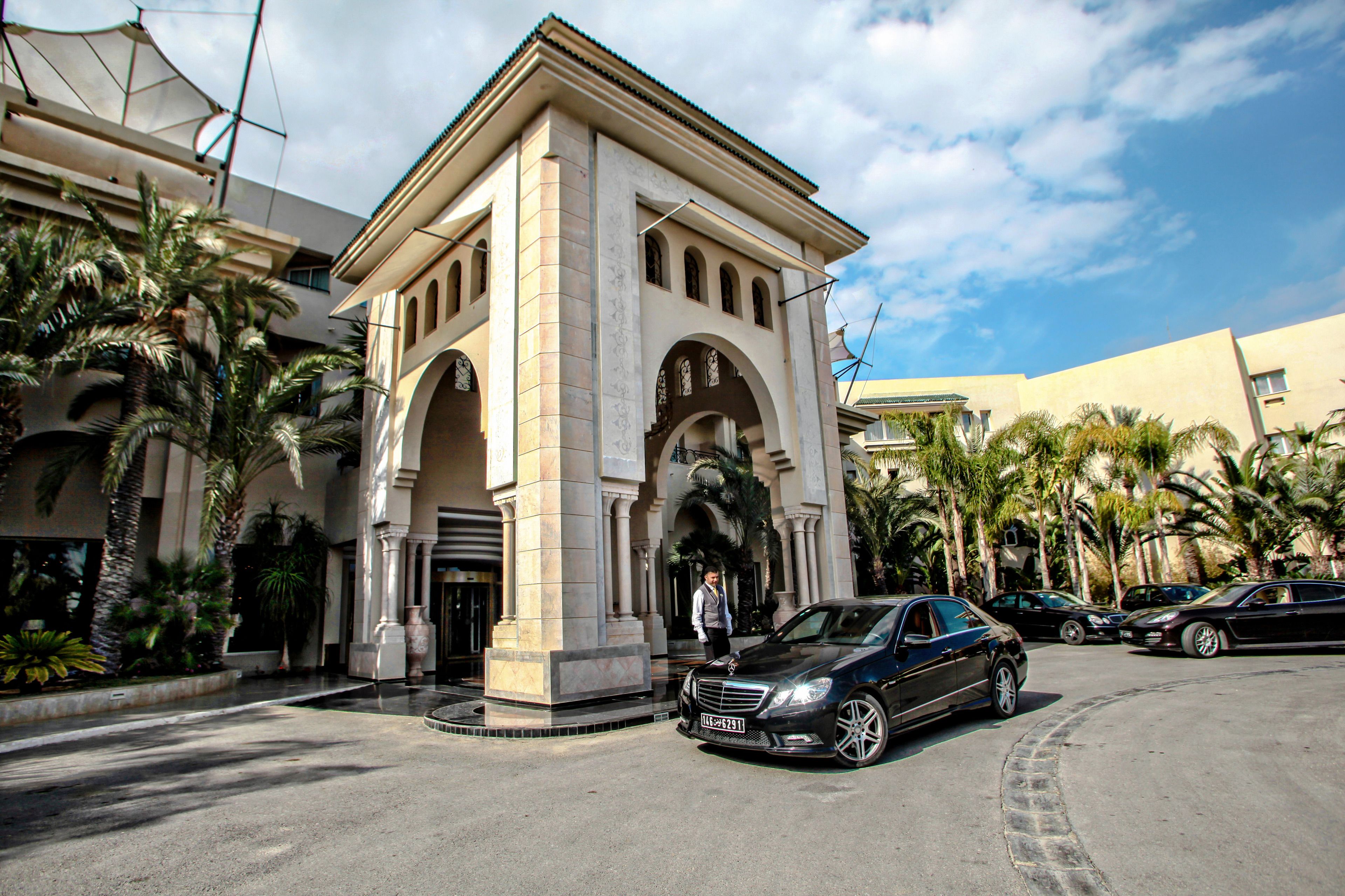 Un edificio con una entrada central en arco, palmeras y coches estacionados.