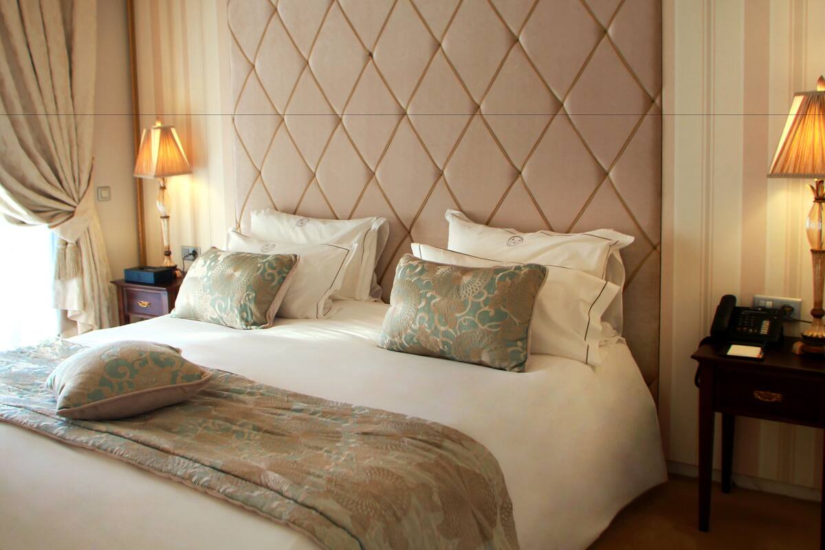 A neatly made bed with a patterned bedspread and pillows, a tufted headboard, bedside lamps, and a nightstand with a telephone.