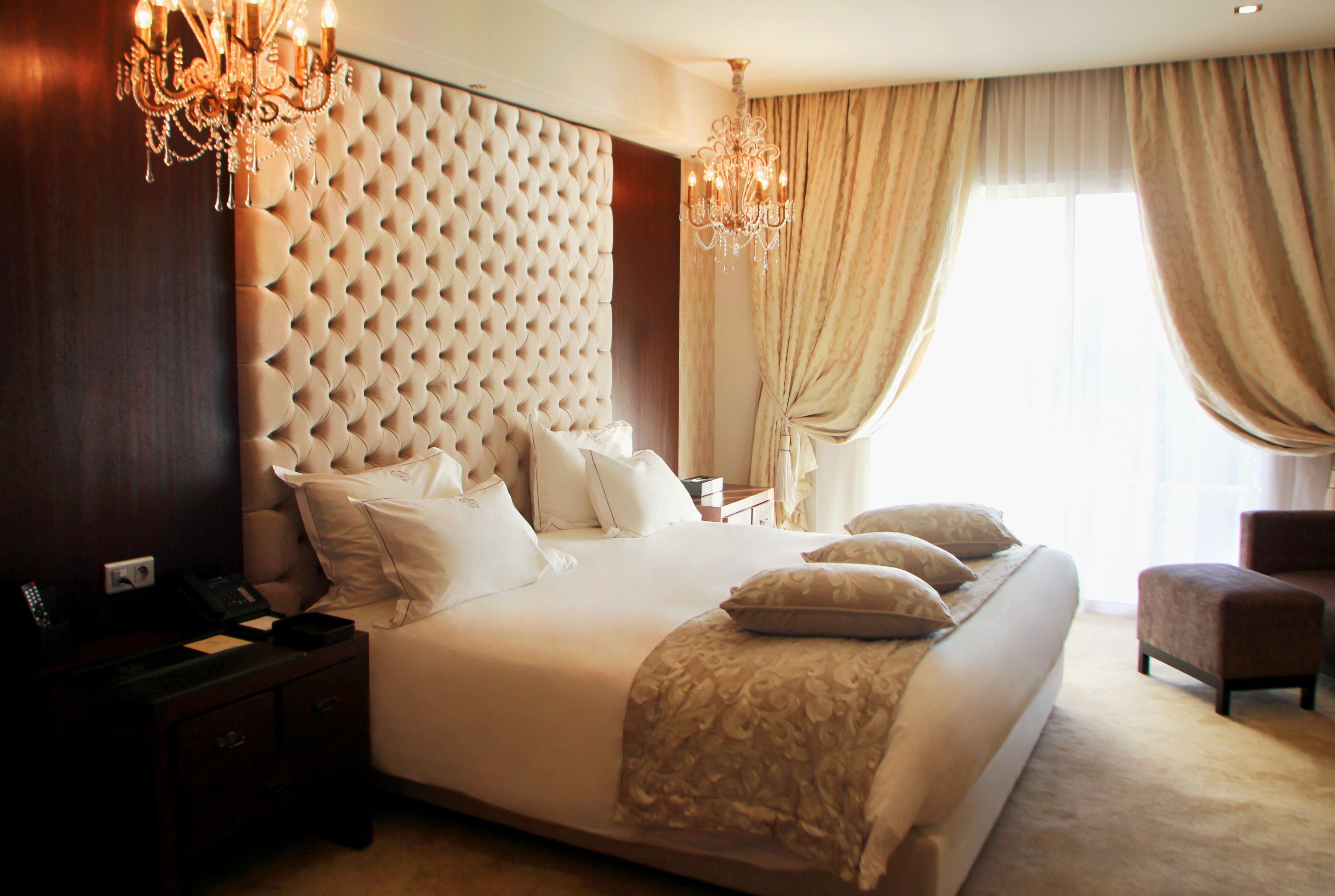 A large bed with a tufted headboard, a bedside table with a phone, and a chair in a well-lit room with a chandelier.
