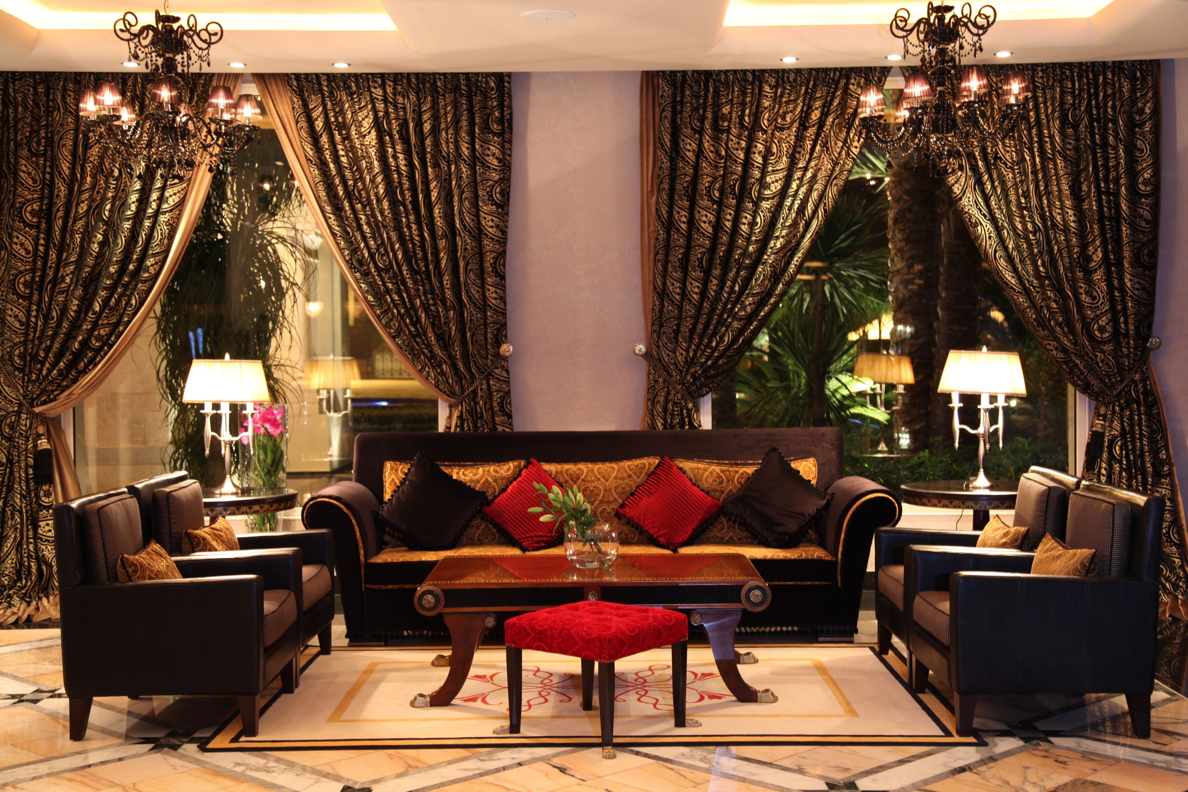 A luxurious living room with a sofa, two armchairs, a coffee table, and a red ottoman. There are two lamps on side tables, a vase with flowers, and curtains with a paisley pattern.