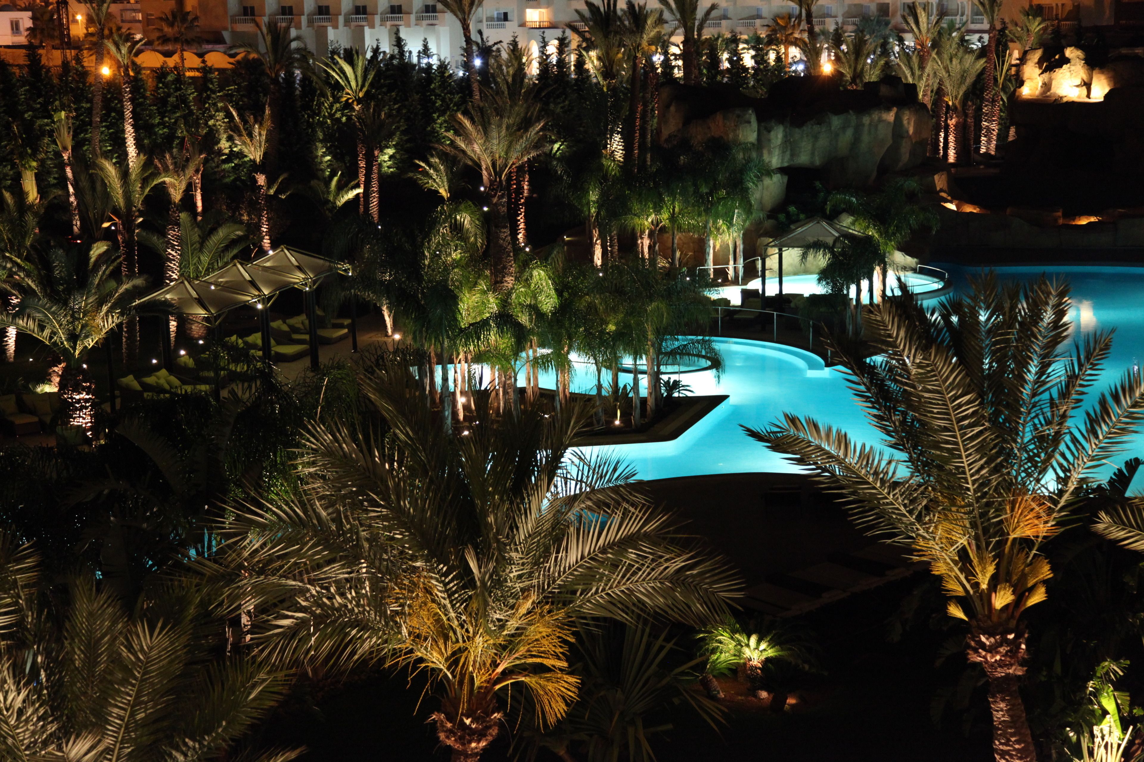 Ein Pool in einem Resort bei Nacht, umgeben von Palmen und einem Gebäude mit Balkonen.