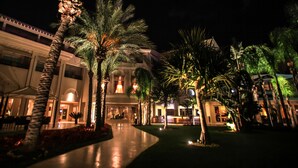 Vista nocturna de un patio de hotel con palmeras, un área de estar cubierta y la entrada del edificio.