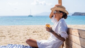 A person sitting on a wooden bench at a beach, holding a glass of beer. - Dionysos Beach Resort & Villas (Ios)