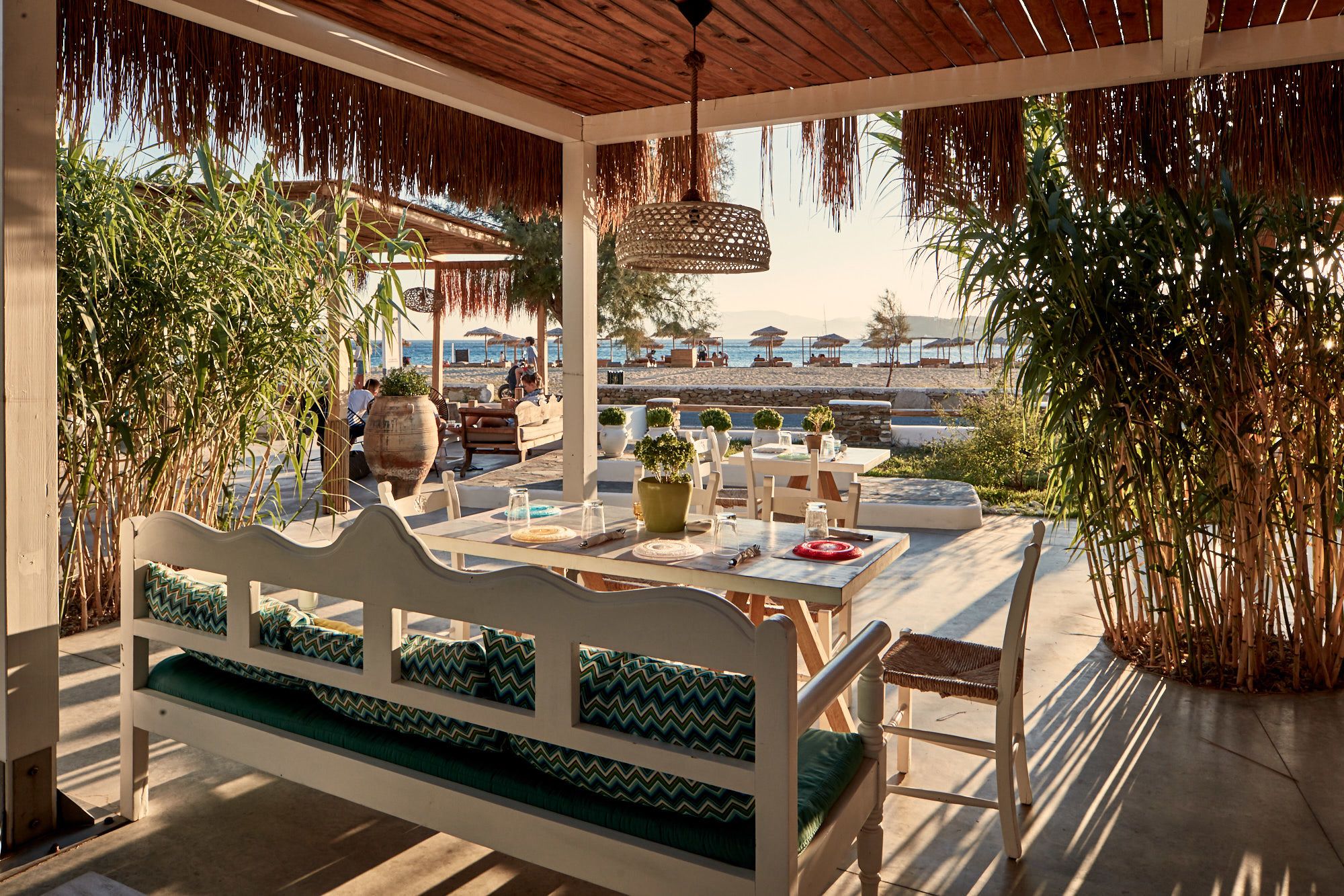 An outdoor seating area with a wooden bench and tables, surrounded by greenery and overlooking a beach.