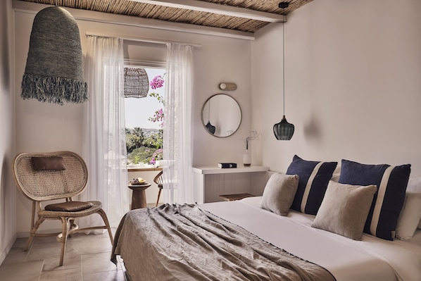 A bedroom with a bed, a chair, a bedside table, a window with curtains, and a hanging light fixture. - Dionysos Beach Resort & Villas (Ios)
