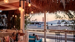 A beachside bar with hanging light bulbs, a bar counter with glasses and bottles, and a person relaxing on a blue couch. - Dionysos Beach Resort & Villas (Ios)