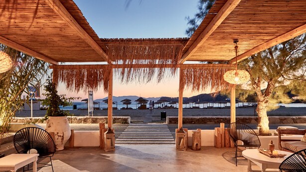 An outdoor seating area with wooden tables and chairs, a thatched roof, and a view of the beach and mountains in the background. - Dionysos Beach Resort & Villas (Ios)